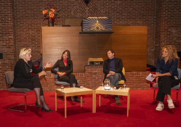 Vier mensen zitten op een stoel naast elkaar met twee tafeltjes voor zich en voeren een gesprek.