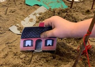 Zandbak waarin een kleuter een papieren huisje vasthoudt