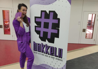 Een mevrouw in een paars pak staat naast een banner van #bakkeln