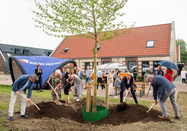 Foto van een groep mensen met scheppen die een boom hebben geplant en het modder er nu weer bij scheppen.