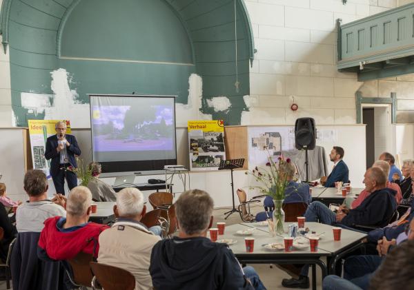 Afbeelding van een groep mensen in een kerk die luisteren naar wethouder Prins