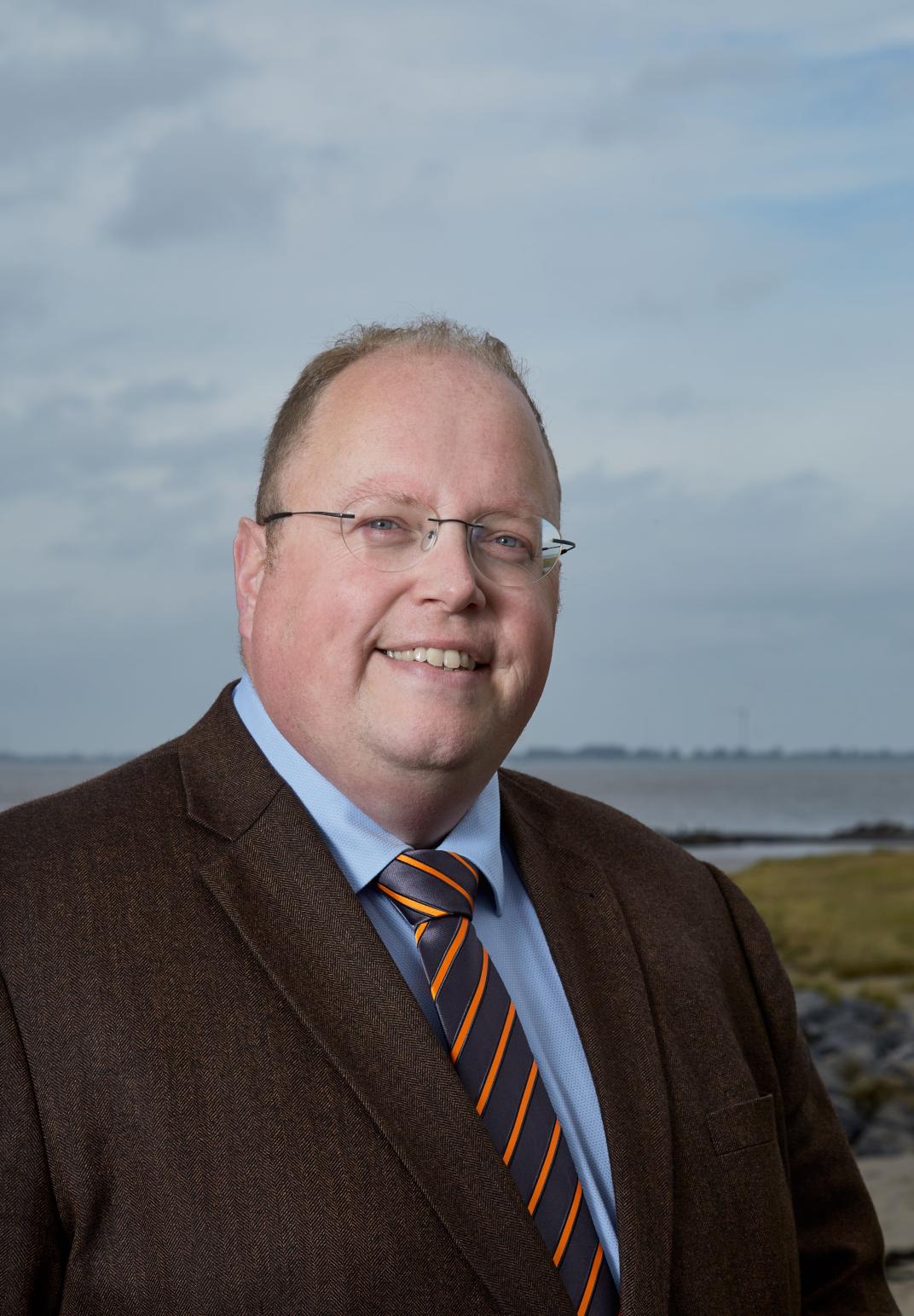 Burgemeester Ben Visser kijkt lachend in de camera, op de achtergrond water, lucht en een stukje gras