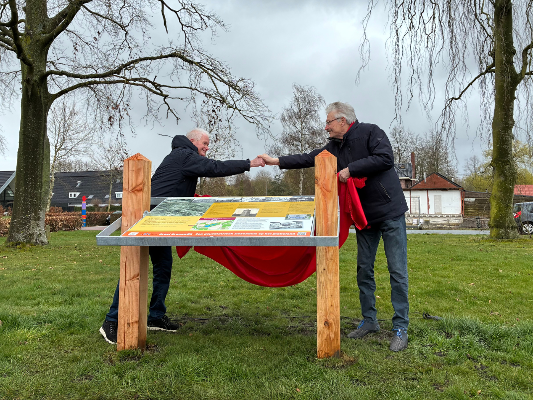 2 personen schudden elkaar de hand boven een soort informatiepaneel die buiten staat op het grasveld
